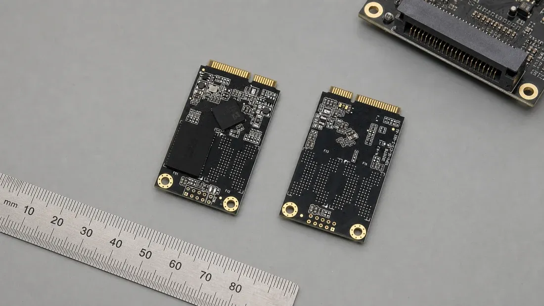 Front and back of two full-size black mSATA SSD modules beside a metric ruler and matching host socket